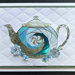 May include: A handmade greeting card featuring a paper teapot design. The teapot is crafted from various shades of blue and teal paper, with a textured, patterned appearance. The card includes light blue paper flowers with silver accents and a white background with a quilted pattern.