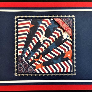 May include: A patriotic greeting card with a red, white, and blue color scheme. The card features a central design of layered paper strips in star and chevron patterns, framed by a silver border and set against a navy blue background.
