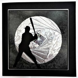 May include: A framed artwork featuring a black silhouette of a baseball player mid-swing. The player is positioned in front of a silver, geometric design resembling a baseball. The background is a dark gray with a subtle floral pattern.