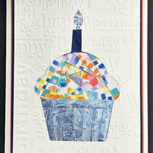 May include: A handmade birthday card featuring a colorful cupcake illustration. The cupcake has a mosaic design with a blue candle and a shimmering flame. The card has embossed "happy birthday" text and a blue and orange border.
