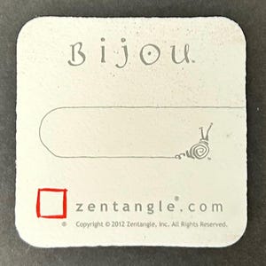 May include: A white, square Bijou Zentangle tag with rounded corners. The tag features the word "Bijou" in a whimsical font, a line for a string, and a snail illustration. The website zentangle.com is printed at the bottom.
