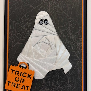 May include: A rectangular greeting card featuring a white, glittery, paper-pieced mummy character with googly eyes. The mummy holds an orange paper bag with the black text TRICK OR TREAT. The background is black with a white spiderweb pattern.