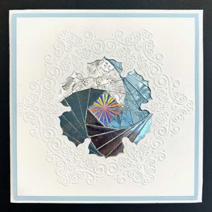 May include: A square greeting card with a light blue border and a white embossed frame featuring intricate scrollwork. The center displays a multi-layered, fan-shaped iris folding design in shades of silver, blue, and black with a holographic center.