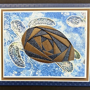 May include: A handmade greeting card featuring a sea turtle design. The turtle's shell is a mosaic of brown and navy blue geometric shapes. The background has a blue watercolor effect with turtle silhouettes.