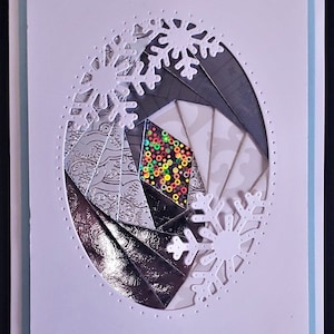 May include: A handmade greeting card with a white background and a light blue border. The card features a cut-out oval design with a geometric pattern of silver, gray, and patterned paper. White snowflake cutouts are placed around the oval.