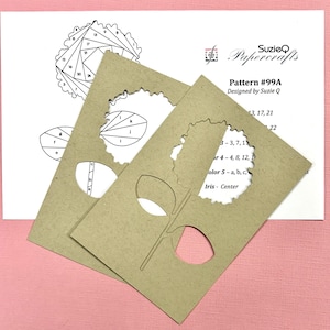 May include: Two tan paper craft stencils with flower and leaf cutouts are layered over a white paper with a floral diagram and text. The text reads "SuzieQ Papercrafts" and "Pattern #99A Designed by Suzie Q".