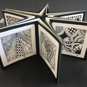 May include: A star-shaped, open book with black and white patterned pages. Each page features a unique, intricate design in black ink on a white background. The book is bound with a spiral spine and a black and white ribbon.