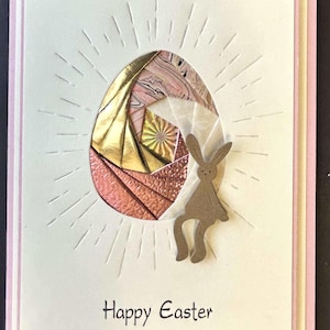 May include: Easter greeting card with a paper-crafted egg design featuring gold, pink, and copper tones. A brown bunny silhouette sits beside the egg. The words "Happy Easter" are printed at the bottom. The card has a pink border.