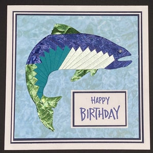May include: A handmade birthday card featuring a stylized fish design. The fish is composed of textured blue, green, and white paper. The card includes the words "Happy Birthday" in blue lettering on a white rectangle.