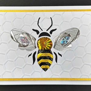 May include: A decorative greeting card featuring a stylized bee design. The bee has silver wings with iridescent accents, a gold and black body, and black antennae. The background is white with a honeycomb pattern, framed by a gold border.