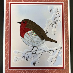 May include: A greeting card featuring a papercraft robin perched on a snow-covered branch. The bird has a red breast, brown wings, and a black beak. The card has a red and white dotted border.