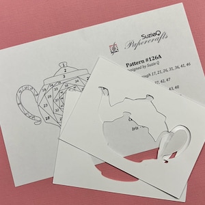 May include: A papercraft pattern by Suzie Q, featuring a teapot design. The pattern includes a diagram with numbered sections and a cut-out template. The background is a light pink color.