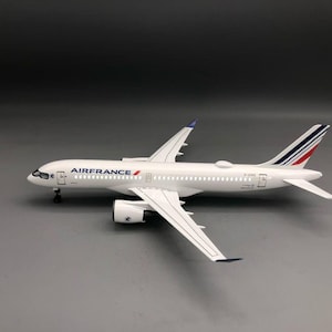 Könnte beinhalten: Ein weißes Air France Modellflugzeug mit einem blau-weiß-roten gestreiften Seitenleitwerk. Der Rumpf zeigt das Air France Logo in blauer Schrift mit einem roten Akzent. Das Flugzeug hat zwei Strahltriebwerke unter den Flügeln und ein Fahrwerk.