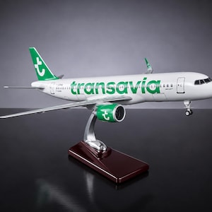 Peut inclure: Un modèle réduit d'avion présentant un fuselage blanc avec le logo et le texte Transavia en vert. L'appareil possède des capots moteurs verts avec un logo blanc, une dérive verte avec un logo blanc, monté sur un socle rectangulaire brun foncé.