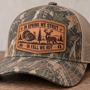 Puede incluir: Una gorra de béisbol de camuflaje con parte trasera de malla color canela y un parche de cuero marrón en el frente. El parche presenta un grabado de un pavo y un ciervo con el texto IN SPRING WE STRUT y IN FALL WE RUT, junto con huellas.