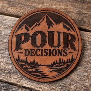 May include: A circular brown leather coaster featuring a laser-engraved design of mountain peaks above a forest and a river. The text "POUR DECISIONS" is centered in bold, dark lettering within the circular border of the coaster.