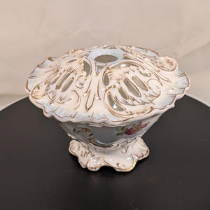 May include: A porcelain flower frog bowl featuring a light blue glaze, gold-painted scalloped edges, and intricate cut-out patterns on the lid. The base is decorated with small pink floral motifs and gold accents, standing on a scalloped pedestal.