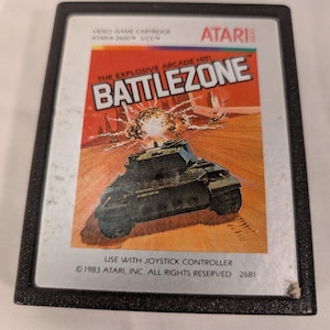 May include: A black Atari 2600 video game cartridge for Battlezone. The label features a green tank in a desert landscape with an explosion, the text "THE EXPLOSIVE ARCADE HIT! BATTLEZONE", "VIDEO GAME CARTRIDGE", and "USE WITH JOYSTICK CONTROLLER".