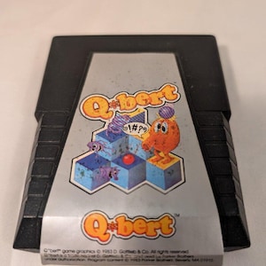 Peut inclure: Une cartouche de jeu vidéo Atari 2600 noire pour Q*bert. L'étiquette présente le personnage orange Q*bert sur une pyramide bleue isométrique, avec le titre du jeu en texte jaune et orange. Le copyright de 1983 de Gottlieb & Co. est en bas.