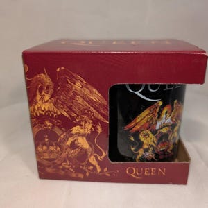 May include: A black ceramic mug featuring the Queen band logo and crest, housed in a maroon cardboard display box. The box displays the word QUEEN in gold lettering at the top and bottom, with a matching gold crest graphic on the front panel.