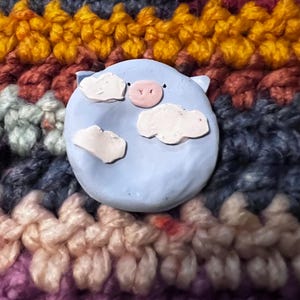 May include: A round, light blue clay ornament featuring a small pink snout and three white, textured cloud shapes attached to the surface. The ornament has two small black dots for eyes and is positioned against a multi-colored, chunky knit background.
