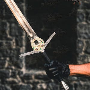 May include: A metal sword featuring a long blade with etched runic inscriptions. The crossguard is circular and gold-toned, accented with small red and green gemstones. The handle is wrapped in black material, ending in a silver-toned pommel.