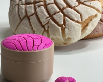 Puede incluir: Un recipiente cilíndrico de color canela con una tapa rosa brillante que presenta un patrón texturizado similar a una concha, acompañado de dos pequeños botones circulares a juego en rosa y canela, frente a un pan dulce redondo y beige.
