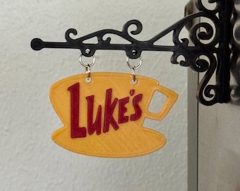 Puede incluir: Un letrero amarillo con forma de taza y el texto rojo "Luke's" colgado de un soporte de metal negro ornamentado con volutas. El letrero está suspendido por dos pequeños anillos de metal plateado unidos a la parte superior de la base amarilla.
