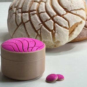 Puede incluir: Un recipiente redondo de color tostado con una tapa rosa brillante que presenta un patr&oacute;n texturizado similar a una concha. Junto al recipiente hay dos peque&ntilde;os objetos circulares a juego en rosa y tostado, con un pan dulce concha al fondo.