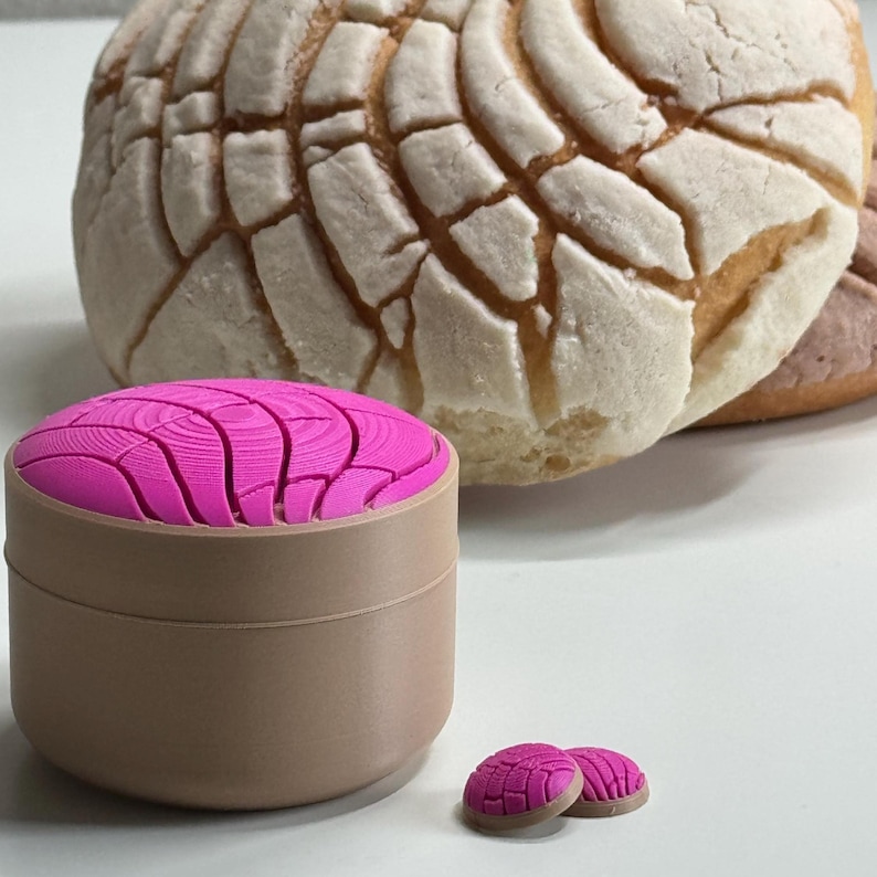 Puede incluir: Un recipiente redondo de color tostado con una tapa rosa brillante que presenta un patr&oacute;n texturizado similar a una concha. Junto al recipiente hay dos peque&ntilde;os objetos circulares a juego en rosa y tostado, con un pan dulce concha al fondo.