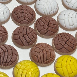 Puede incluir: Una colecci&oacute;n de adornos redondos en forma de pan concha con un patr&oacute;n texturizado de concha en la parte superior. El conjunto incluye piezas blancas, marrones y de color amarillo brillante, cada una con una base plana y lisa y una superficie detallada.