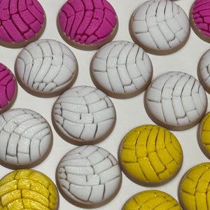 Puede incluir: Una colecci&oacute;n de adornos redondos con textura de pan concha en rosa brillante, blanco y amarillo. Cada pieza presenta un patr&oacute;n distintivo similar a una concha en la parte superior con una base lisa de color marr&oacute;n claro, dispuestos en cuadr&iacute;cula.