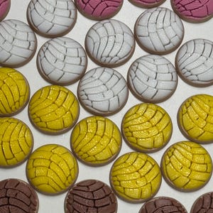 Puede incluir: Una colecci&oacute;n de adornos redondos en forma de pan concha en colores rosa, blanco, amarillo y marr&oacute;n. Cada pieza presenta un patr&oacute;n texturizado similar a una concha en la parte superior, imitando el aspecto del pan dulce mexicano.