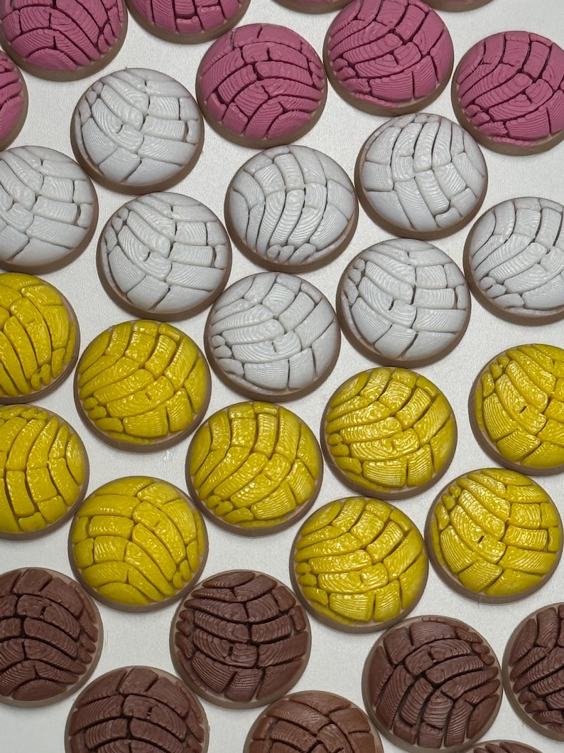 Puede incluir: Una colecci&oacute;n de adornos redondos en forma de pan concha en colores rosa, blanco, amarillo y marr&oacute;n. Cada pieza presenta un patr&oacute;n texturizado similar a una concha en la parte superior, imitando el aspecto del pan dulce mexicano.
