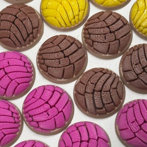 Puede incluir: Una colecci&oacute;n de adornos redondos en forma de c&uacute;pula que representan pan concha en colores marr&oacute;n, amarillo brillante y rosa intenso. Cada pieza presenta un patr&oacute;n texturizado similar a una concha en la parte superior, imitando el pan dulce.