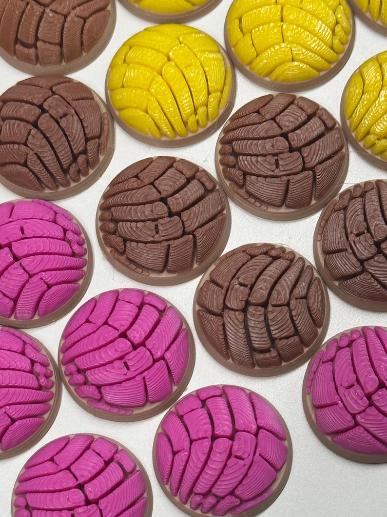 Puede incluir: Una colecci&oacute;n de adornos redondos en forma de c&uacute;pula que representan pan concha en colores marr&oacute;n, amarillo brillante y rosa intenso. Cada pieza presenta un patr&oacute;n texturizado similar a una concha en la parte superior, imitando el pan dulce.