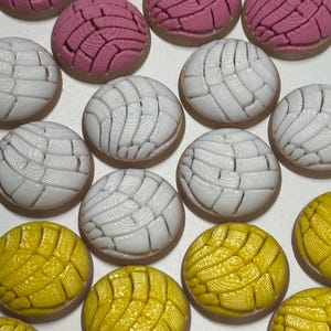 Puede incluir: Una colecci&oacute;n de adornos redondos con forma de pan concha en rosa, blanco y amarillo. Cada pieza presenta un patr&oacute;n texturizado similar a una concha en la parte superior con una base lisa de color marr&oacute;n claro, dispuestos en cuadr&iacute;cula.