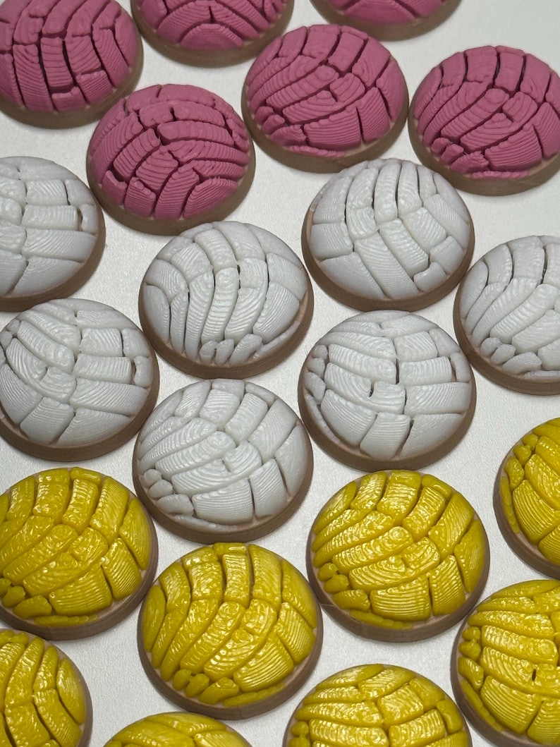 Puede incluir: Una colecci&oacute;n de adornos redondos con forma de pan concha en rosa, blanco y amarillo. Cada pieza presenta un patr&oacute;n texturizado similar a una concha en la parte superior con una base lisa de color marr&oacute;n claro, dispuestos en cuadr&iacute;cula.