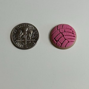 Puede incluir: Un peque&ntilde;o dije redondo de pan concha rosa con un patr&oacute;n texturizado similar a una concha en la parte superior y una base color canela, colocado junto a una moneda de diez centavos de EE. UU. para escala, que mide aproximadamente 1,79 cm de di&aacute;metro.