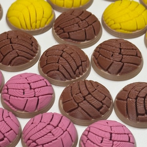 Puede incluir: Una colecci&oacute;n de adornos redondos y texturizados de pan concha en amarillo, marr&oacute;n y rosa. Cada pieza presenta un patr&oacute;n distintivo en forma de concha en la superficie superior, imitando la apariencia de los panecillos dulces mexicanos.