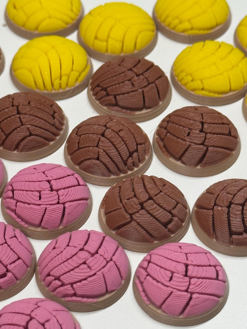 Puede incluir: Una colecci&oacute;n de adornos redondos y texturizados de pan concha en amarillo, marr&oacute;n y rosa. Cada pieza presenta un patr&oacute;n distintivo en forma de concha en la superficie superior, imitando la apariencia de los panecillos dulces mexicanos.