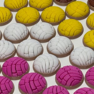 Puede incluir: Una colecci&oacute;n de miniaturas de pan concha, peque&ntilde;as, redondas y en forma de c&uacute;pula, en colores amarillo brillante, blanco y rosa vibrante. Cada pieza presenta un patr&oacute;n texturizado en forma de concha en la parte superior, imitando el pan dulce.