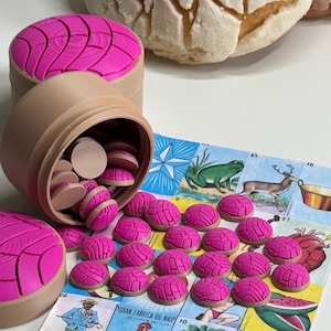 Puede incluir: Un recipiente cil&iacute;ndrico de color canela con tapa, lleno y rodeado de peque&ntilde;as fichas redondas con forma de concha y parte superior rosa. Las fichas presentan un patr&oacute;n texturizado similar a una concha en sus superficies rosas sobre un tablero.