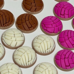 Puede incluir: Una colecci&oacute;n de adornos redondos en forma de pan concha en colores marr&oacute;n, rosa brillante y blanco. Cada pieza presenta un patr&oacute;n texturizado similar a una concha en la parte superior, imitando el aspecto de los panecillos dulces.