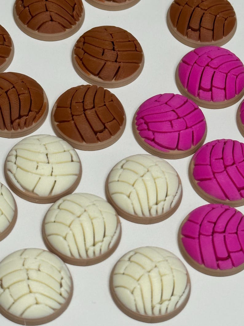 Puede incluir: Una colecci&oacute;n de adornos redondos en forma de pan concha en colores marr&oacute;n, rosa brillante y blanco. Cada pieza presenta un patr&oacute;n texturizado similar a una concha en la parte superior, imitando el aspecto de los panecillos dulces.