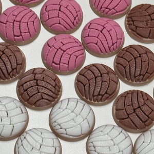 Puede incluir: Una colecci&oacute;n de adornos redondos en forma de pan concha en colores rosa, marr&oacute;n y blanco. Cada pieza presenta un patr&oacute;n texturizado similar a una concha en la superficie superior, imitando el aspecto de los panecillos dulces.