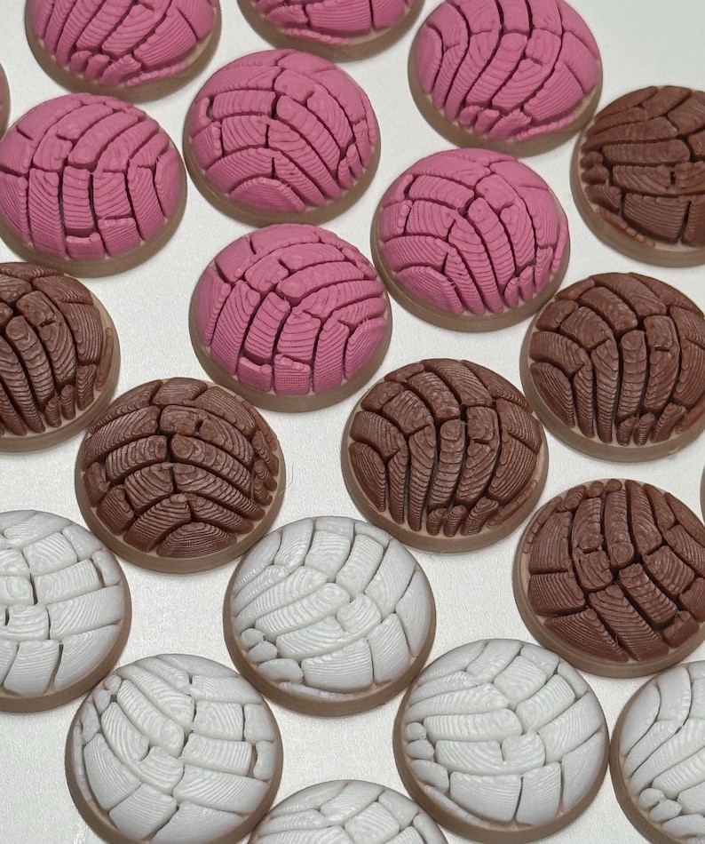 Puede incluir: Una colecci&oacute;n de adornos redondos en forma de pan concha en colores rosa, marr&oacute;n y blanco. Cada pieza presenta un patr&oacute;n texturizado similar a una concha en la superficie superior, imitando el aspecto de los panecillos dulces.