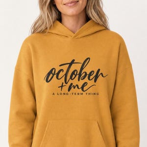May include: A mustard yellow hooded sweatshirt featuring the black text "october + me" in a cursive script font, with the additional text "A LONG-TERM THING" printed in a smaller, sans-serif font directly underneath the main graphic.