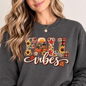 May include: A charcoal grey crewneck sweatshirt featuring a graphic print with the text FALL VIBES. The letters are filled with illustrations of sunflowers, orange pumpkins, red maple leaves, acorns, and a plaid pattern against a sunset landscape.