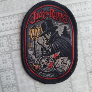 May include: An oval embroidered patch featuring the text Jack the Ripper in red. The design depicts a figure in a top hat and coat holding a magnifying glass, a street lamp, a smoking pipe, a bloody knife, and a paper labelled CLUES in grey and black.